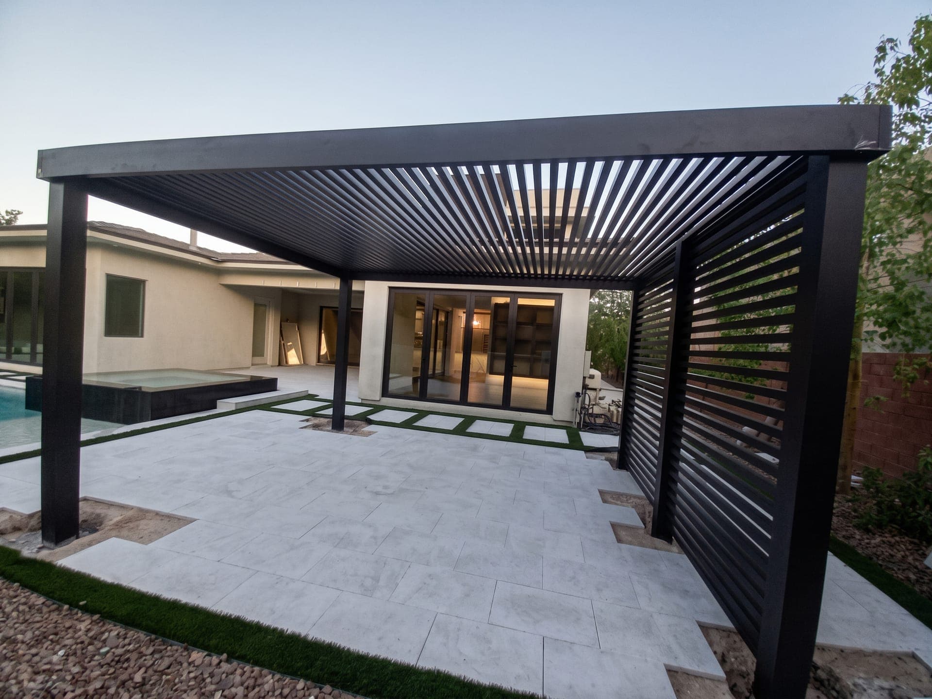 Dark 4K Aluminum freestanding lattice pergola with a horizontal slat privacy wall beside a pool and spa at dusk