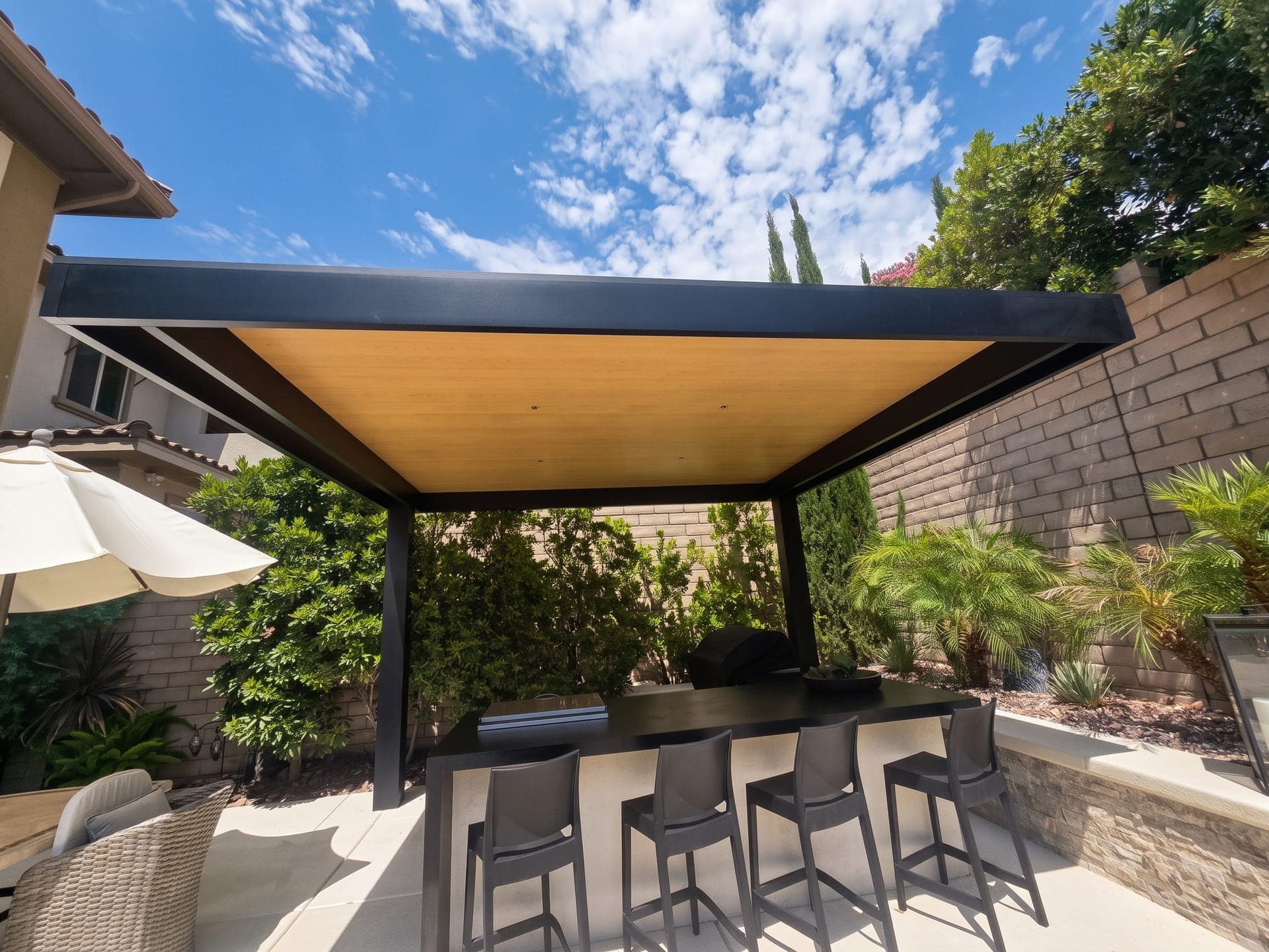 4K Aluminum cantilever cover with a solid roof, dark frame and wood-look ceiling over an outdoor bar in a walled backyard